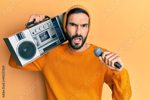 Young hispanic man holding boombox, listening to music singing with microphone in shock face, looking skeptical and sarcastic, surprised with open mouth