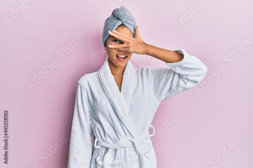 Young chinese woman wearing shower towel cap and bathrobe peeking in shock covering face and eyes with hand, looking through fingers with embarrassed expression.