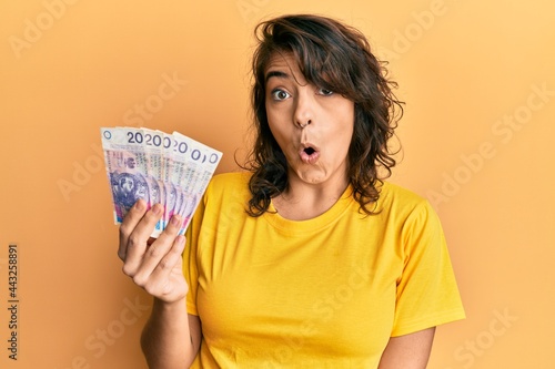 Young hispanic woman holding 20 polish zloty banknotes scared and amazed with open mouth for surprise, disbelief face