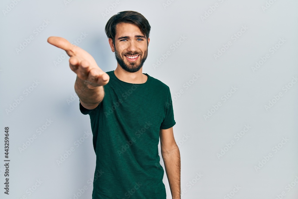 Fototapeta premium Young hispanic man wearing casual white tshirt smiling friendly offering handshake as greeting and welcoming. successful business.