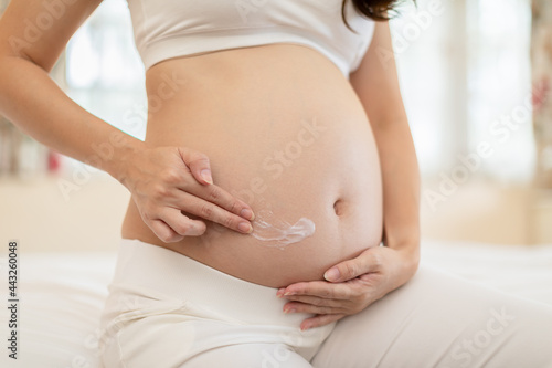 Happy Pregnant Woman sitting on bed holding and stroking apply cream on big belly for beauty moisturizing skin at home,Pregnancy young woman enjoying with future life,Motherhood and Pregnant Concept