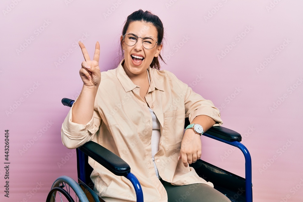Young hispanic woman sitting on wheelchair smiling with happy face winking at the camera doing victory sign with fingers. number two. Young hispanic woman sitting on wheelchair smiling with happy face winking at the camera doing victory sign with fingers. number two.