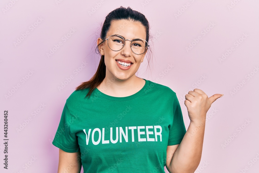 Young hispanic woman wearing volunteer t shirt pointing to the back behind with hand and thumbs up, smiling confident Young hispanic woman wearing volunteer t shirt pointing to the back behind with hand and thumbs up, smiling confident