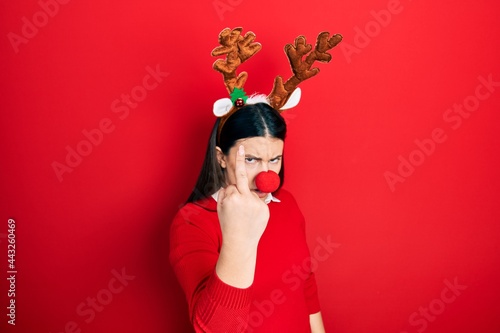 Young hispanic woman wearing deer christmas hat and red nose showing middle finger, impolite and rude fuck off expression