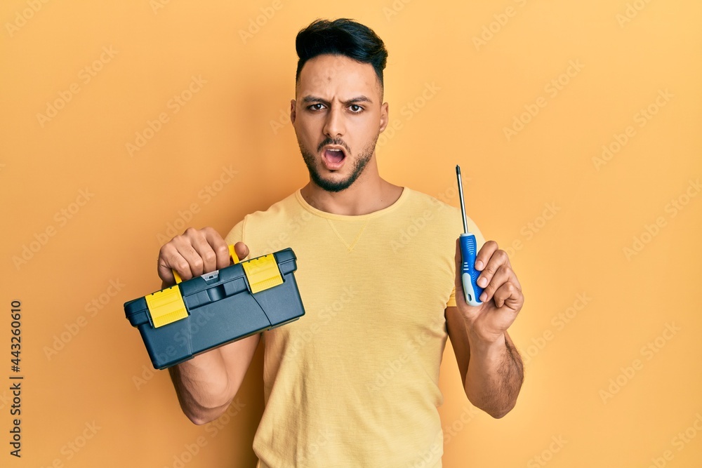 Young arab man holding toolbox and screwdriver in shock face, looking skeptical and sarcastic, surprised with open mouth Young arab man holding toolbox and screwdriver in shock face, looking skeptical and sarcastic, surprised with open mouth