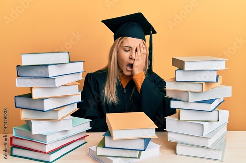 Young caucasian woman wearing graduation ceremony robe sitting on the table yawning tired covering half face, eye and mouth with hand. face hurts in pain.
