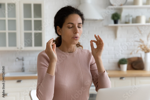 Mindful young woman breathing out with closed eyes, calming down in stressful situation, working on computer in modern kitchen. Millennial hispanic lady managing stress, practice yoga at home office.