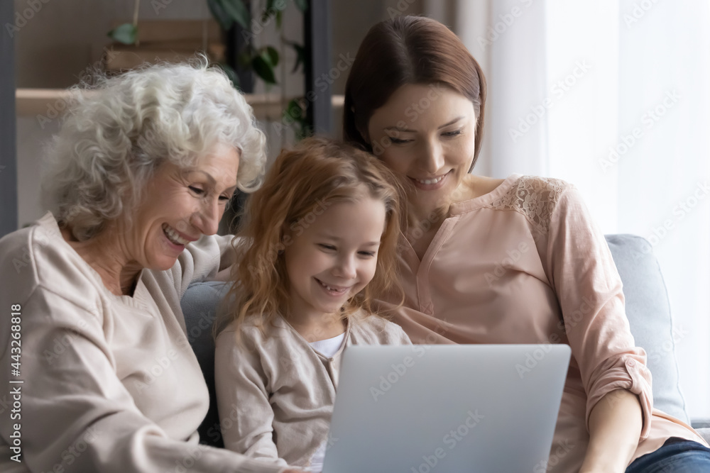 Three age family. Happy little girl daughter grandkid sit on sofa between elderly grandma and adult mom watch video movie on laptop. Smiling females of diverse generations surf internet on pc together Three age family. Happy little girl daughter grandkid sit on sofa between elderly grandma and adult mom watch video movie on laptop. Smiling females of diverse generations surf internet on pc together