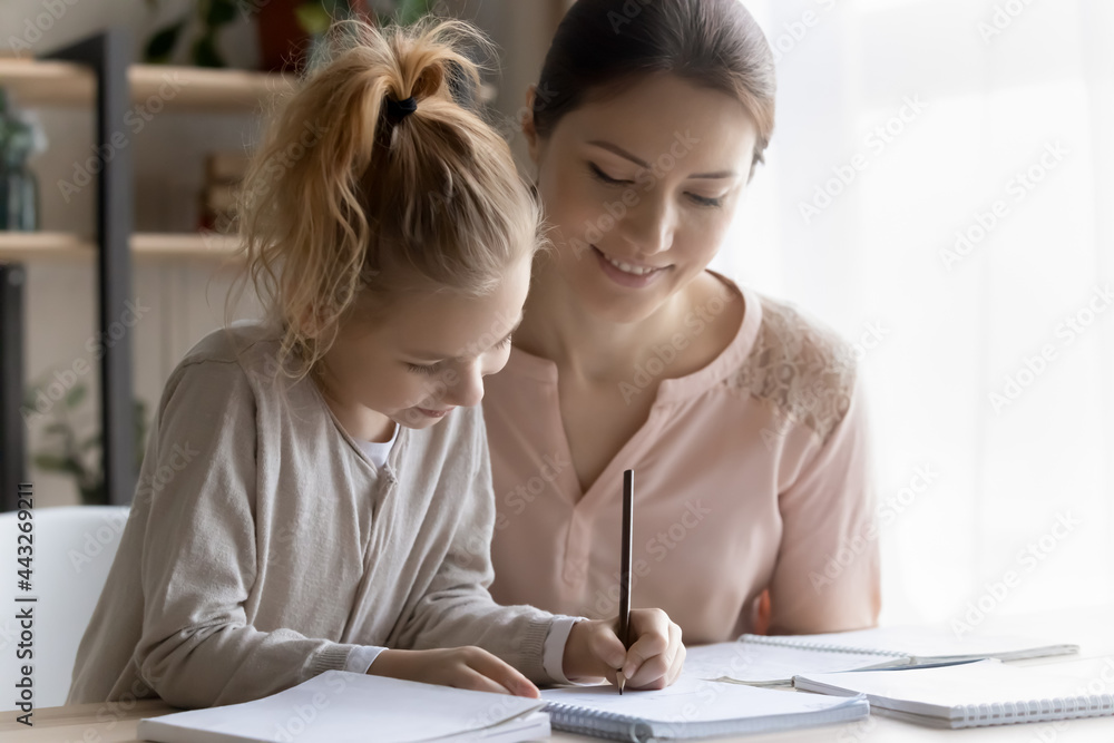 Fototapeta premium Out of school education. Helping young mom engaged in learning activity with preteen daughter assist in studying maths or english rules. Smiling millennial lady teacher give home lesson to little girl