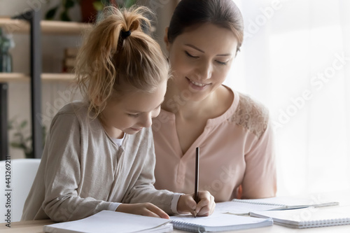 Out of school education. Helping young mom engaged in learning activity with preteen daughter assist in studying maths or english rules. Smiling millennial lady teacher give home lesson to little girl