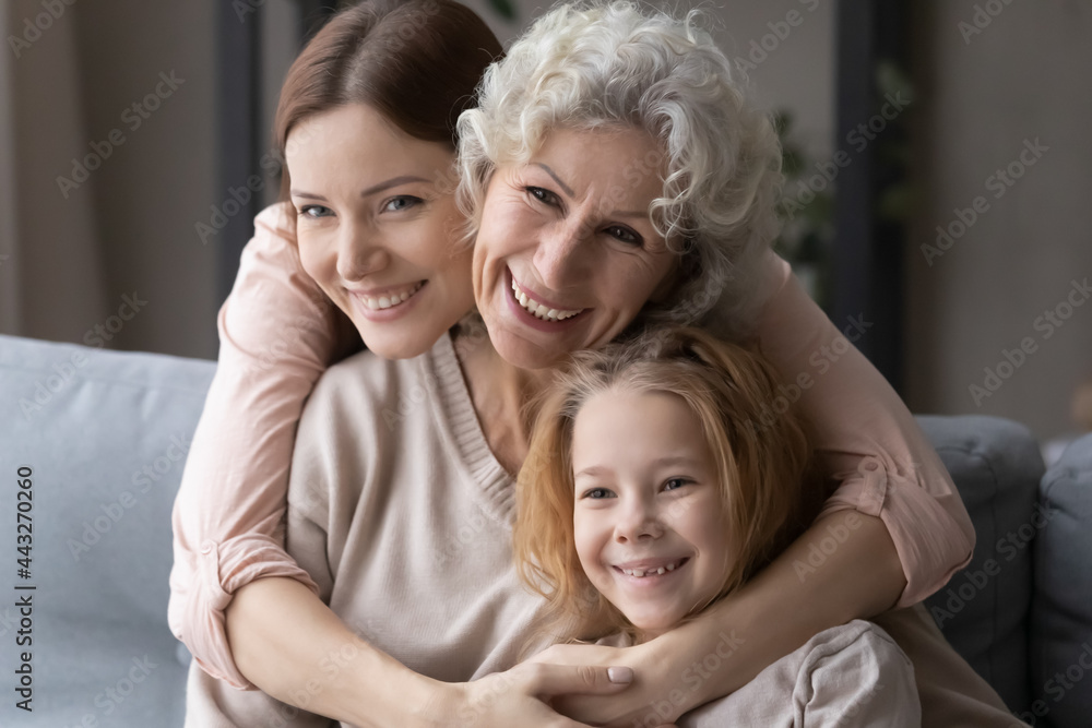 Portrait of 3 generations family. Happy young female embrace from back smiling mature mother and preteen child girl. Caring grey haired grandma sit on sofa cuddle little grandkid and adult daughter