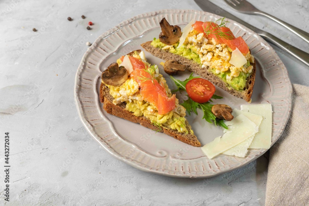 Two sandwiches with scrambled eggs, guacamole, smoked salmon and vegetables on rye bread. On a gray background, horizontal, with space. Two sandwiches with scrambled eggs, guacamole, smoked salmon and vegetables on rye bread. On a gray background, horizontal, with space.