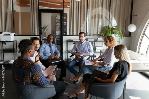 Serious diverse businesspeople discussing project statistics, holding documents, sitting in circle in modern office room, successful employees team colleagues brainstorming financial report