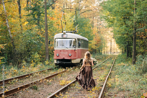A girl in an evening brown dress with a bouquet of flowers goes to meet the tram in the forest photo
