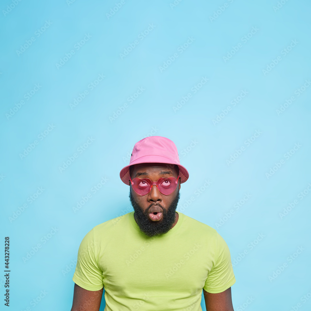 Impressed surprised dark skinned guy with thick beard focused above keeps mouth opened wears panama casaul green t shirt heart shaped sunglasses isolated over blue background with blank space Impressed surprised dark skinned guy with thick beard focused above keeps mouth opened wears panama casaul green t shirt heart shaped sunglasses isolated over blue background with blank space