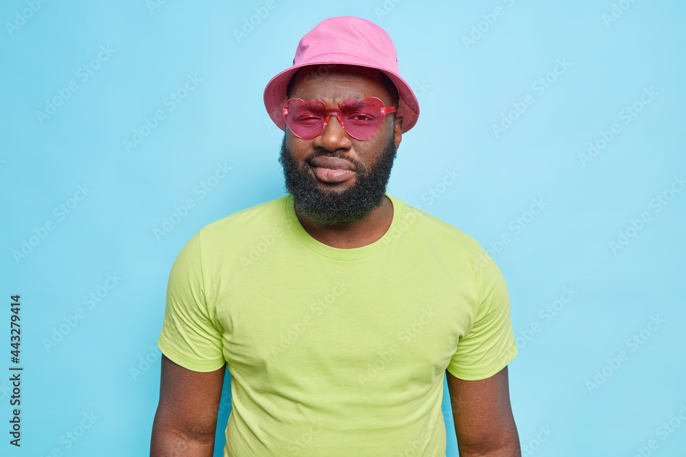 Serious bearded man looks directly at camera wears trendy sunglasses hat and casual green t shirt isolated over blue background. Fashionable hipster guy indoor. People style clothing concept Serious bearded man looks directly at camera wears trendy sunglasses hat and casual green t shirt isolated over blue background. Fashionable hipster guy indoor. People style clothing concept