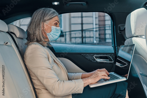 Mature beautiful woman in protective face mask using laptop while sitting on the back car seat