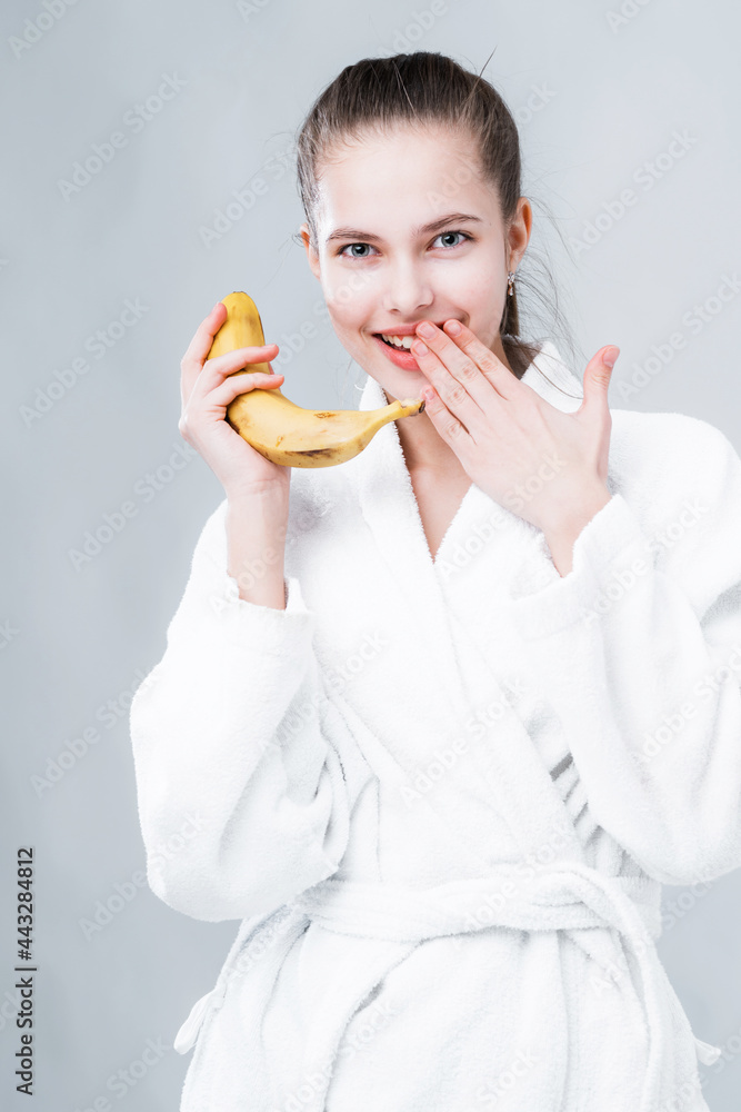 Attractive girl in a bathrobe holds a banana in her hand and smiles covering her mouth with her hand