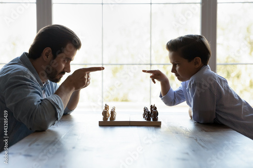 Funny severe dad and little son playing chess, beginning game, sitting opposite like competitors, opponent players at chessboard with start sets, changing comical threatening challenging looks photo