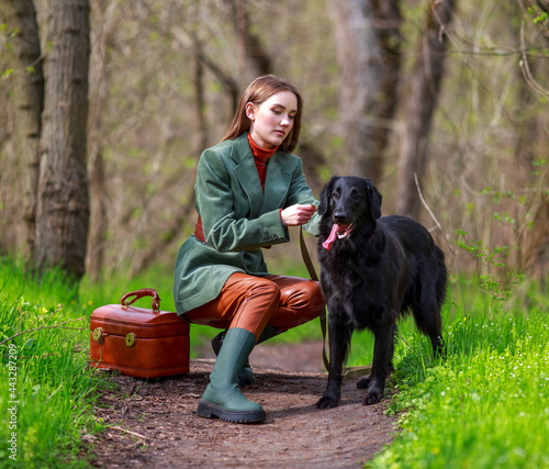 Girl in stylish rustic country clothes with black retriever hunting dog walking in green forest