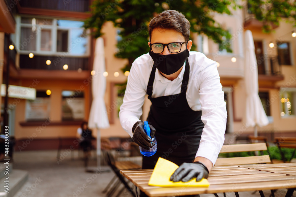 Disinfecting to prevent COVID-19. Waiter in protective face mask and gloves cleaning table with disinfectant spray and Microfiber cloth in cafe in quarantine city. Disinfecting to prevent COVID-19. Waiter in protective face mask and gloves cleaning table with disinfectant spray and Microfiber cloth in cafe in quarantine city.