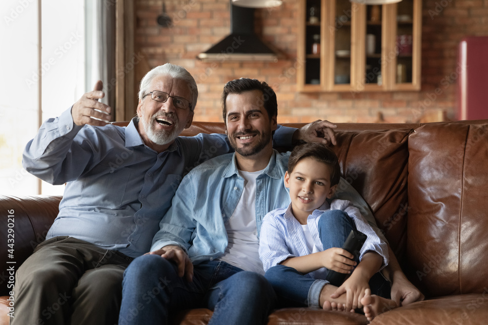 Happy preschooler boy, young dad, excited mature senior 70s grandpa watching TV in living room, sitting on couch together at home, holding remote control. Leisure with family, three generations