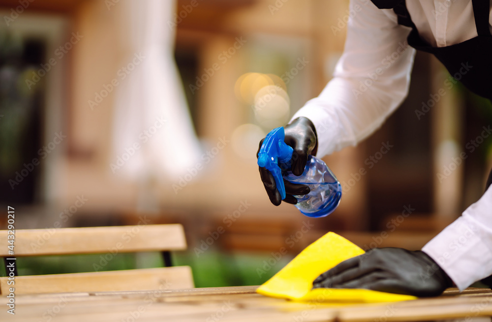 Disinfecting to prevent COVID-19. Waiter in protective face mask and gloves cleaning table with disinfectant spray and Microfiber cloth in cafe  in quarantine city.