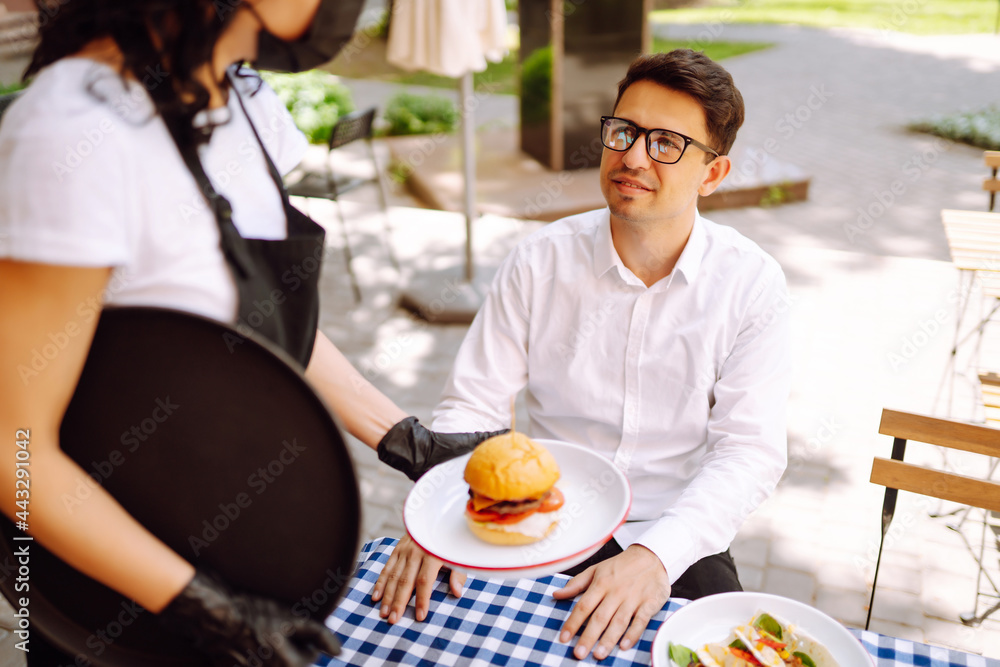 Young waitress in a protective medical mask and gloves with ordered meals serves food at table outside. Coronavirus outbreak lifestyle. Social distancing. Young waitress in a protective medical mask and gloves with ordered meals serves food at table outside. Coronavirus outbreak lifestyle. Social distancing.