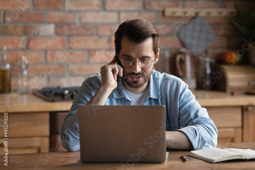 Serious millennial remote employee working at laptop from home, talking to client on mobile phone, answering customer, giving consultation, support. Man using computer and making call at kitchen table