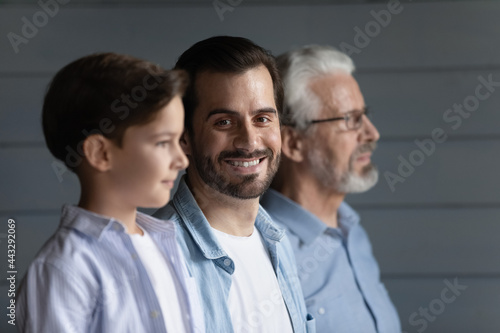 Happy young millennial father standing in row with son boy and mature 70s grandfather, looking at camera, smiling. Family portrait of three male generations. Intergenerational relations concept