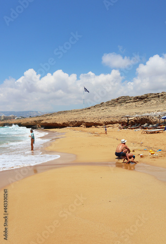Yellow sandy beach near coral bay with turquoise blue water, white foamy waves, beachgoers and the Greek flag in the distance