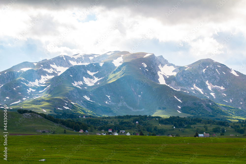 A view of a huge mountain range at the foot of which are houses. Snow can be seen on the mountain and there is beautiful green grass at the foot.
