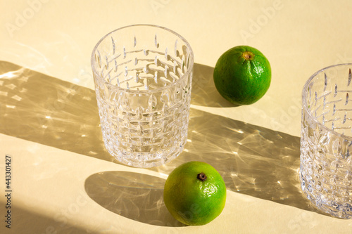 a line of empty crystal glasses and two limes under hard sunlight, glitter reflection on beige background, long harsh rectangular and oval shadows