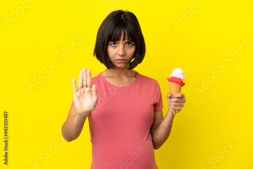 Pregnant woman holding a cornet ice cream isolated on yellow background making stop gesture © luismolinero
