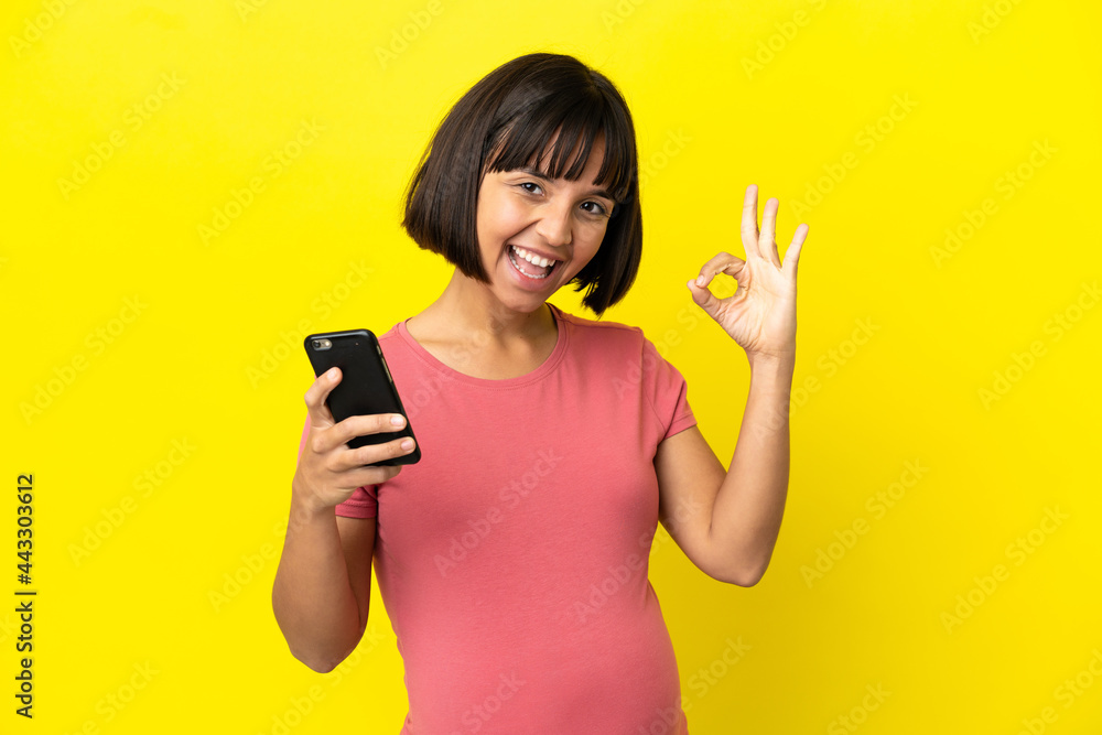 Young mixed race pregnant woman isolated on yellow background using mobile phone and doing OK sign Young mixed race pregnant woman isolated on yellow background using mobile phone and doing OK sign