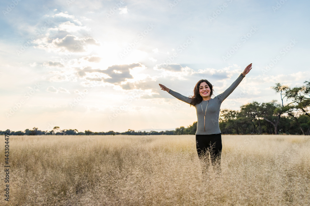 Young adult Mexican smiling woman standing with open arms in a dry grassy meadow Young adult Mexican smiling woman standing with open arms in a dry grassy meadow