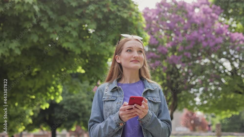 Portrait of happy young Caucasian beautiful female walking in park and texting on smartphone. Close up of joyful smiling woman tapping on mobile phone outdoors and looking away. Slow motion. Portrait of happy young Caucasian beautiful female walking in park and texting on smartphone. Close up of joyful smiling woman tapping on mobile phone outdoors and looking away. Slow motion.
