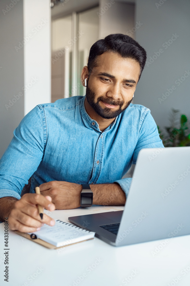 Portrait of handsome young indian man student watching online webinar using laptop and earphone making noes in notebook. Video conference call, business training tutorial, coaching course concept Portrait of handsome young indian man student watching online webinar using laptop and earphone making noes in notebook. Video conference call, business training tutorial, coaching course concept