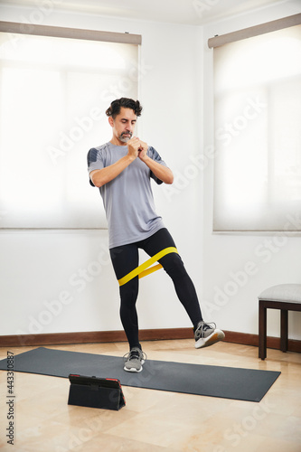 Middle aged man with beard doing exercise at home with rubber band looking video on tablet