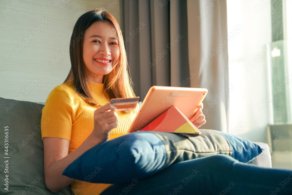 Asian woman sitting on sofa using digital tablet with credit card for online shopping. Female shopper enjoy purchase goods in online store with e-banking online payment on mobile application at home.