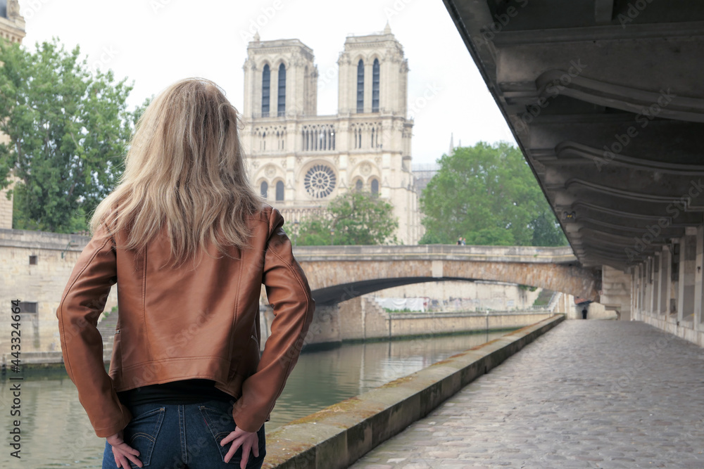 Seductive blonde woman in trousers, standing from behind on the banks of the Seine river. Cathedral of Notre Dame de Paris in the background.