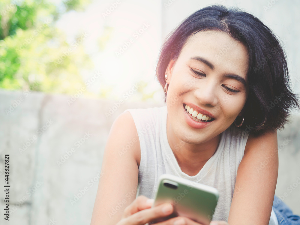 Happy beautiful Asian woman with short hair in casual white sleeveless shirt laughing while looking her mobile phone on green nature background with copy space. Happy beautiful Asian woman with short hair in casual white sleeveless shirt laughing while looking her mobile phone on green nature background with copy space.