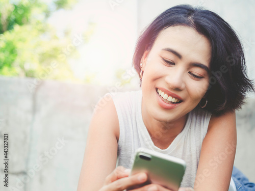 Happy beautiful Asian woman with short hair in casual white sleeveless shirt laughing while looking her mobile phone on green nature background with copy space.