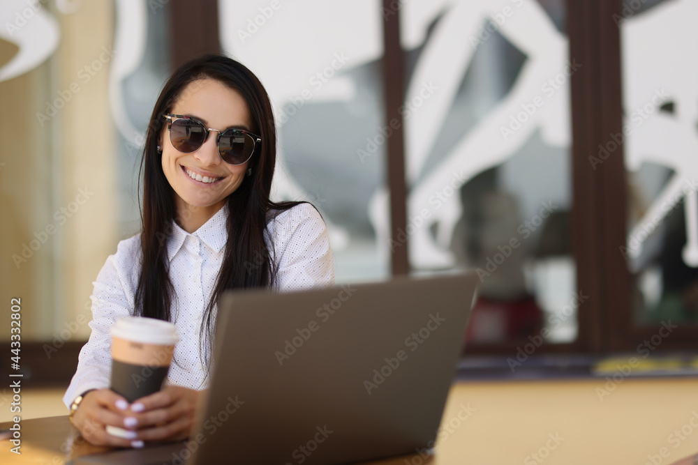 Young woman in sunglasses sitting at table with laptop and drinking coffee on cafe terrace