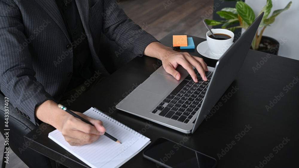 Cropped shot businessman making notion notebook and using laptop at office desk. Cropped shot businessman making notion notebook and using laptop at office desk.