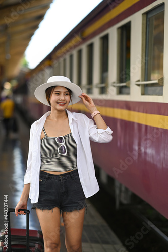 Cheerful woman traveler wearing white hat walking with her suitcase at platform.