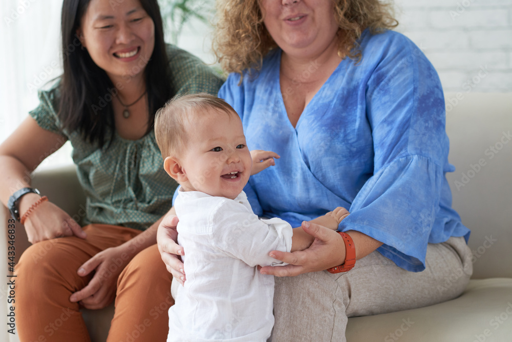Mother sitting on sofa next to girlfriend and supporting her little baby making first steps Mother sitting on sofa next to girlfriend and supporting her little baby making first steps