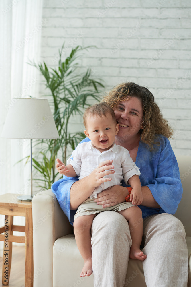Portrait of happy smiling mother sitting on sofa with little baby on her laps when spending weekend at home Portrait of happy smiling mother sitting on sofa with little baby on her laps when spending weekend at home