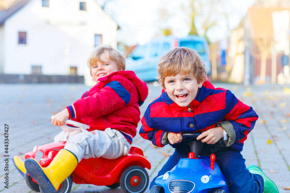 Obraz premium Two little kids boys in colorful clothes and rain boots driving toy cars. Siblings making competition, outdoors. Active leisure for children on autumn day.