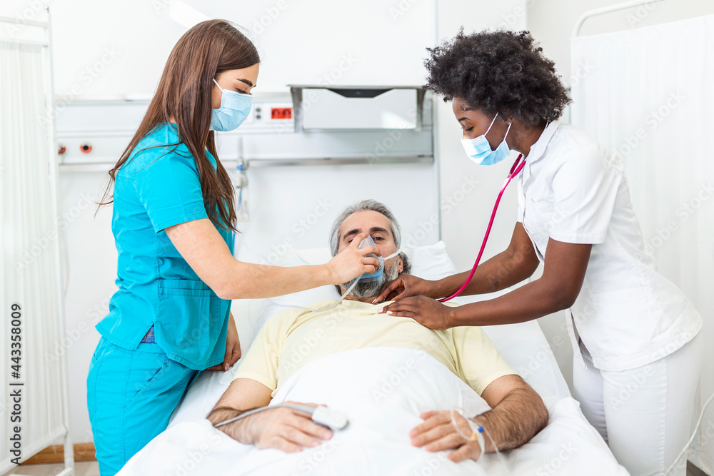 Nurse puts oxygen mask on elderly man patient lying in the hospital room bed while African American doctor examining patient with stethoscope, coronavirus covid 19 protection concept Nurse puts oxygen mask on elderly man patient lying in the hospital room bed while African American doctor examining patient with stethoscope, coronavirus covid 19 protection concept